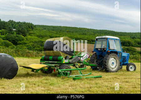 Ballydehob, West Cork, Irlanda. Il 25 giugno, 2018. Alan Pyburn di Durrus basato John O'Driscoll silaggio appaltatori avvolge un insilato bail sul Ballydehob farm di Ben Deane in una giornata molto calda. Questo è il preludio a una mini-ondata di caldo in cui le temperature sono impostati per raggiungere 31° gradi centigradi da giovedì di questa settimana. Credito: Andy Gibson/Alamy Live News. Foto Stock