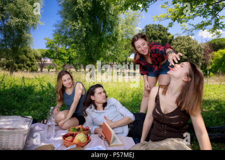 Le ragazze e i ragazzi che giace in erba durante un picnic al parco. Una ragazza offre una fetta di anguria al suo amico, il ragazzo che legge un libro e si mangia. In Foto Stock