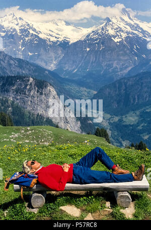 Una femmina di escursionista poggia su una panca in legno convenientemente situato lungo un sentiero che offre vedute panoramiche della Jungfrau e altre vette montane nell Oberland Bernese Regione della Svizzera. L'itinerario a piedi di Schynige Platte plateau si snoda tra fiori selvatici alpini che compaiono in estate dopo la neve si scioglie in altitudini più elevate delle alpi svizzere. Foto Stock