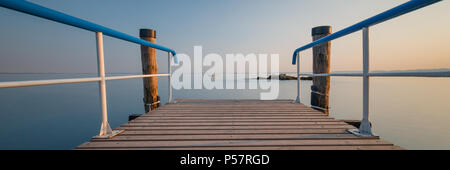 Esposizione di lungo periodo di un molo in legno con ringhiera blu su una soleggiata sera in riva al lago di Garda, Italia Foto Stock