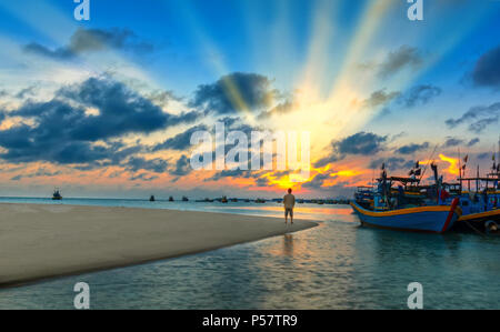 La Lonely Man orologi il tramonto su una spiaggia di sabbia vicino al villaggio di pescatori con tante barche ancorate in acque lontane Foto Stock