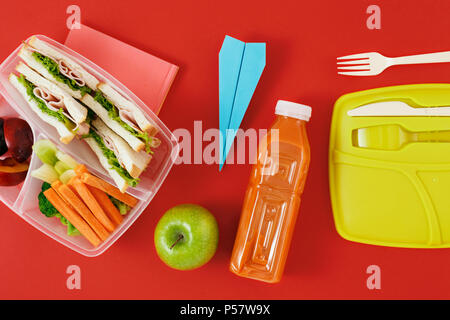 Pranzo sano di scatole con sandwich e verdure fresche, bottiglia di succo di frutta e carta piano su sfondo rosso. vista dall'alto. Cibo sano concetto. Lay piatto co Foto Stock