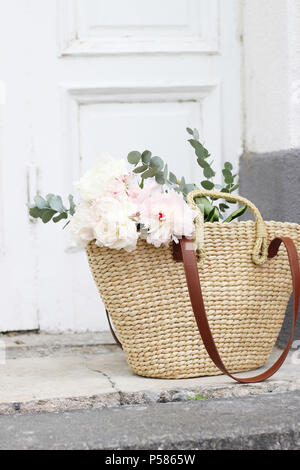 Elegante stock photo. Nozze femminile ancora vita composizione con paglia francese basket borsa con peonie rosa fiori e bouquet di eucalipto. Vecchia porta bianca in background. Composizione verticale. Foto Stock