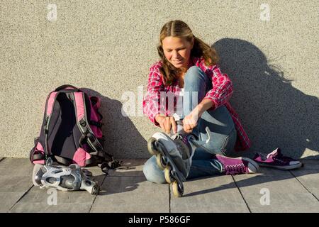 Active sport teen gir in quad pattini a rotelle, background urbano. Ragazza adolescente rimuove sneakers e vestiti pattini a rotelle all'aperto. Si siede su una parete backgr Foto Stock