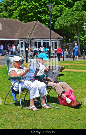 Una coppia di mezz'età rilassante sotto il sole e la lettura di quotidiani sul verde a Christchurch Quay, Dorset.Il Vecchio Mulino sale da tè sono in background. Foto Stock