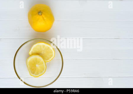 Un limone e due fette di limone nella lastra di vetro.Un limone è sulla parte superiore e due fette di limone nella lastra di vetro sono verso il basso.bianco sullo sfondo di legno.Copys Foto Stock