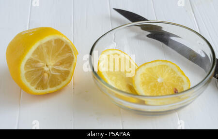 Un limone tagliato e due fette di limone nella lastra di vetro.parte della lama di coltello dietro la lastra di vetro. Bianco sullo sfondo di legno. Foto Stock