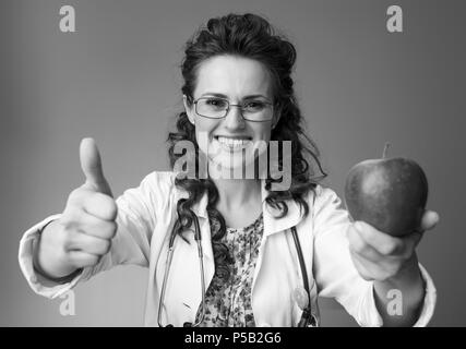 Sorridente medico pediatra in bianco manto medico dando un apple e mostrando pollice in alto sullo sfondo Foto Stock