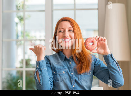 Redhead donna ciambella di contenimento a casa rivolto con la mano e il dito verso l'alto con la faccia felice sorridente Foto Stock