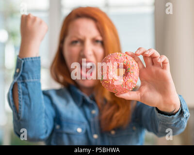 Redhead donna ciambella di contenimento a casa arrabbiato e frustrato grida con rabbia, crazy e urla con la mano alzata, concetto di rabbia Foto Stock