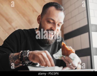 Giovane uomo barbuto mangiare hamburger e sorridendo vicino. Foto Stock