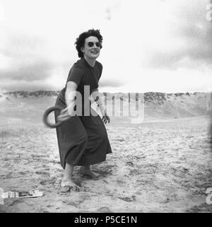Degli anni Cinquanta, donna attraente in occhiali da sole all'aperto su di una spiaggia di sabbia in scalza, indossando un abito giocando a un gioco di gettare il cerchio o l'anello, Inghilterra, Regno Unito. Foto Stock