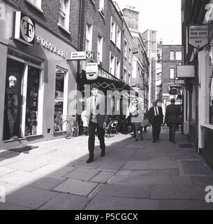 Anni '1960, storia, gente che cammina in un passaggio pavimentato a Shepherd Market, Piccadilly, nell'elegante quartiere di Mayfair a Londra. Edward Shepherd progettò il mercato e le tenute di Grosvenor e Cavendsh. Nella foto si può vedere un negozio della catena di mercanti di vino e liquori Fosters. Fosters, il grande produttore di birra australiano, vendette in seguito i suoi mercanti di vino, che divennero noti come Peter Dominic. Foto Stock