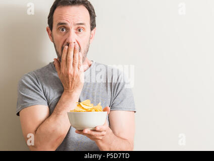 Senior uomo mangiare patatine coprono la bocca con la mano sconvolto con vergogna per errore, espressione di paura, paura in silenzio, concetto segreta Foto Stock