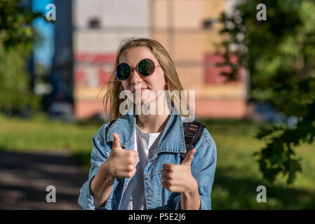 Bella schoolgirl ragazza. In estate nel parco dopo la scuola. In occhiali da sole e in una camicia di denim. Gesto della mano mostra un pollice in su. I cappelli Foto Stock