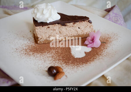 Pezzo di torta al cioccolato con panna montata sulla parte superiore su una lastra decorata con fiore violaceo, mandorle e cosparsi di cioccolato in polvere. Foto Stock