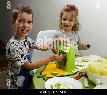 Due bambini piccoli, di due e quattro anni di aiutare a preparare il cibo per il pasto successivo dalla macinazione di zucchine e paprika. Foto Stock