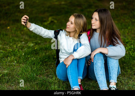 Due ragazze adolescenti. In estate il parco della città. Essi siedono sull'erba e scattare foto sullo smartphone. Dietro gli zaini. Il concetto di scuola di amicizia. Emozione pongono sul telefono. Foto Stock