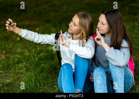 Due ragazze adolescenti. In estate al parco della città. Essi siedono sull'erba e scattare foto con lo smartphone. Dietro gli zaini. Effettuare una chiamata video via Internet. Comunica in reti sociali. Foto Stock