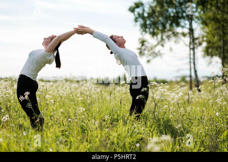Due donne le facendo esercizi yoga, piegatura permanente per ogni altro a all'aperto nel parco naturale. Foto Stock