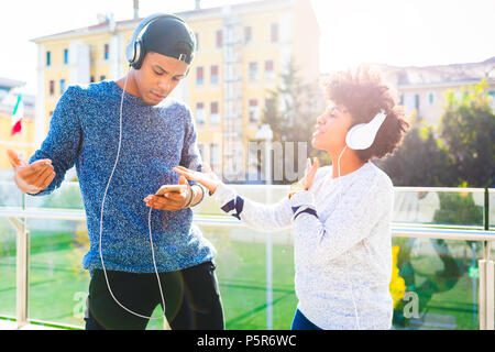 Coppia giovane multirazziale ascoltando la musica ballare outdoor torna la luce utilizzando le cuffie e smart phone - la felicità, tecnologia, concetto di interazione Foto Stock
