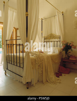 Voile bianco oscuranti su metallo fourposter letto con lenzuola bianche in portoghese camera da letto con pavimento piastrellato di bianco Foto Stock