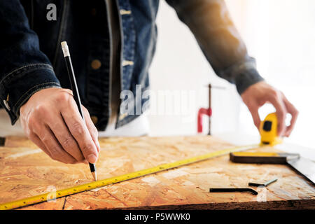Carpenter operando con cautela guardando i piani di lavoro in falegnameria. Egli è imprenditore di successo al suo posto di lavoro. martellare un chiodo supporta sulla build Foto Stock