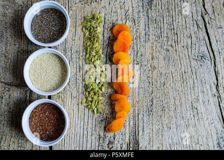 Tre super foods chia, lino e i semi di sesamo in bianco pals su uno sfondo di legno e accanto ai semi di zucca e albicocche secche. Vista dall'alto. Foto Stock