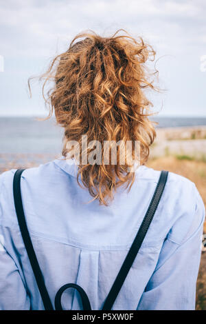 Vista posteriore della giovane donna con spessi capelli ricci in coda di cavallo seduto sul mare. Foto verticale di un irriconoscibile donna indossa maglietta blu e retro Foto Stock