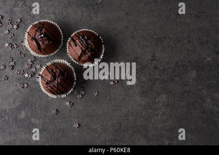 Piatto di laici tre muffin al cioccolato guarnita con cioccolato medicazione circondata con le stelle di cioccolato nero su sfondo di calcestruzzo con copia spazio. Top v Foto Stock