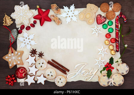 Sfondo di Natale confine con gioia segno compresi addobbi per l'albero, biscotti, dolci, cioccolatini e flora invernale su carta pergamena sul rovere rustico. F Foto Stock