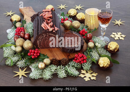 Il cioccolato yule log torta di Natale con sherry, inverno flora, cioccolatini in carta stagnola e decorazioni su rovere rustico sfondo legno. Cibo festoso per h Foto Stock