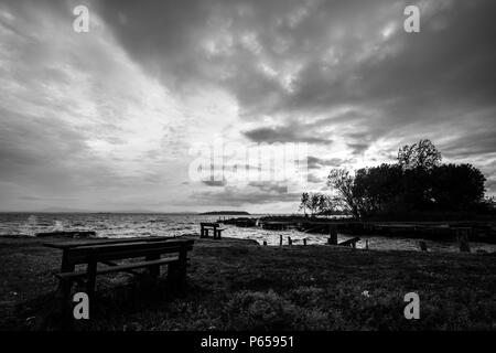 Seduta in legno panchine su un lago al tramonto, al di sotto di un moody, cielo molto nuvoloso Foto Stock