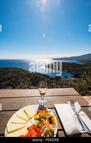 Piastra con insalata di formaggio e accompagnati da una birra e sullo sfondo la baie della la riserva naturale di Cap de Creus sulla Costa Brava in Catalogna Sp Foto Stock