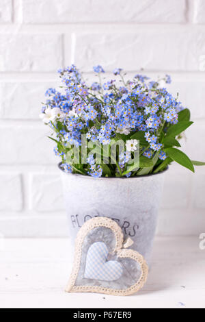 Dimenticare blu-me-poveri o myosotis fiori in grigio la benna e cuore decorativo bianco su sfondo di legno. Floral still life. Messa a fuoco selettiva. Vertic Foto Stock