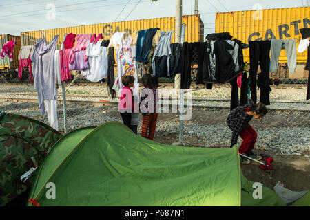 Le ragazze suona insieme accanto alla maglia di filo in cui i vestiti sono essiccazione in un campo di fortuna presso la stazione ferroviaria di frontiera Greek-Macedonian vicino Foto Stock