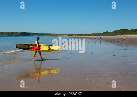 Un bagnino femmina passeggiate dal mare sulla spiaggia di Tenby, Pembrokeshire, Galles portando una scheda di salvataggio Foto Stock