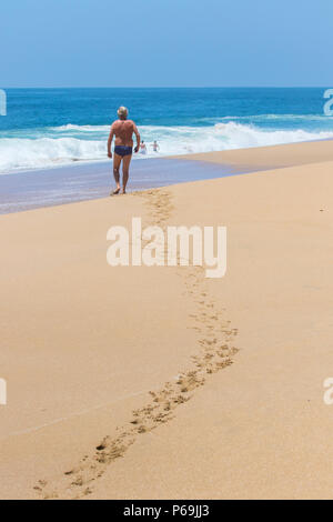 Un Tropicale scena come un anziano uomo che cammina su una spiaggia lascia impronte nella sabbia dorata con un vivido il mare blu e bianco di schiumatura onde in background. Foto Stock