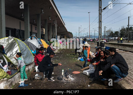 I rifugiati sedi in corrispondenza di un falò tra tende impostato su un sui binari della stazione ferroviaria all'improvvisato campo di rifugiati di frontiera Greek-Macedonian vicino Foto Stock