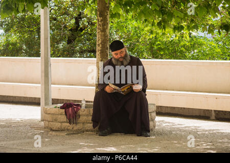 Persone di mezza età barbuto serbo-ortodossa monaco seduto in ombra sotto agli alberi il relax e la lettura di rivista a Ostrog monastero superiore, Danilovgrad, Montenegro Foto Stock