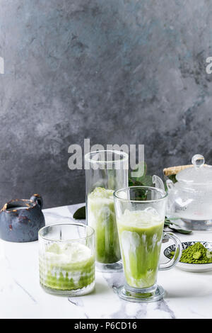 Tè verde Matcha iced latte macchiato o un cocktail in tre diversi tipi di bicchieri con cubetti di ghiaccio, matcha polvere e il contenitore del latte su bianco tavola di marmo, decorata da g Foto Stock
