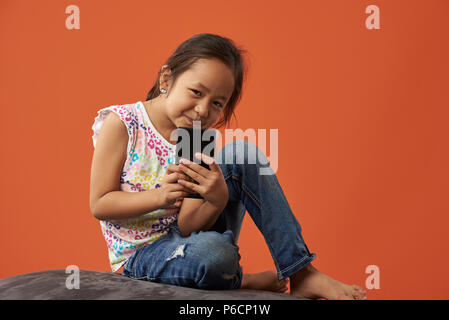 Asian bambina tenere lo smartphone a sedersi su un divano isolato su sfondo arancione Foto Stock