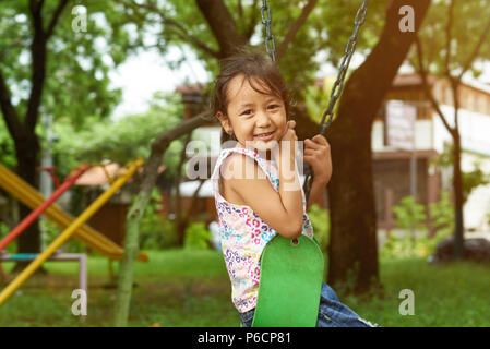 Ritratto di asian piccola ragazza seduta sulle oscillazioni nel soleggiato parco sullo sfondo Foto Stock