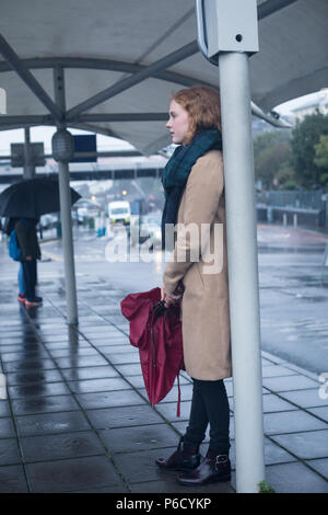 Giovane donna con ombrello in piedi alla fermata del bus Foto Stock