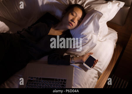 Stanco donna che dorme con il computer portatile e mobile pone sul letto Foto Stock