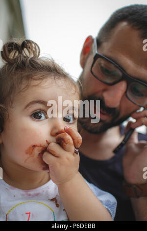 Bambina godendo di una torta al cioccolato mentre suo padre è sul telefono Foto Stock