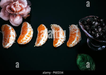 La vitamina C. 5 freschi fette di mandarino e congelate di ribes nero nel piatto di vetro su uno sfondo nero decorato con panno artificiale rosa rosa e verde le Foto Stock