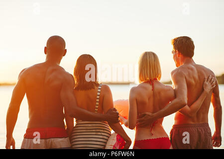 Specchietto di due giovani coppie in costume da bagno in piedi a braccetto insieme in un lago di guardare il tramonto Foto Stock