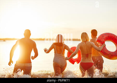 Specchietto di un gruppo di diversi giovani amici in costume da bagno gli spruzzi di acqua durante il funzionamento insieme in un lago al tramonto Foto Stock