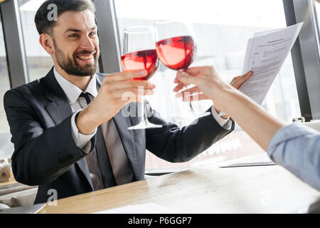 L uomo e la donna avente attività pranzo al ristorante seduti al tavolo vicino a finestra di bere vino rosso cheers celebrando la firma del contratto allegro sorridente Foto Stock